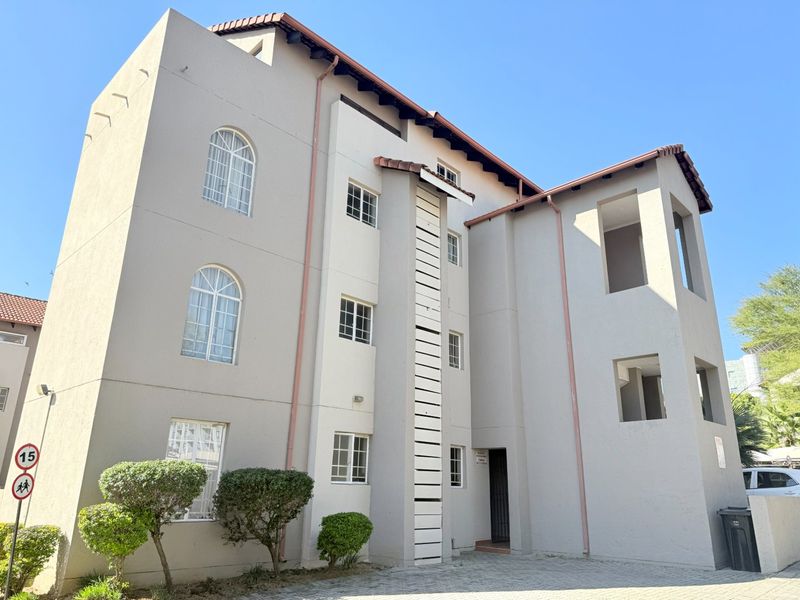 A multi-story apartment building exterior featuring a neutral facade, arched windows, and a paved driveway entrance.