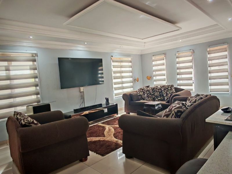 A spacious living room featuring a decorative tray ceiling with recessed lighting, large porcelain floor tiles, and multiple windows fitted with modern roller blinds.