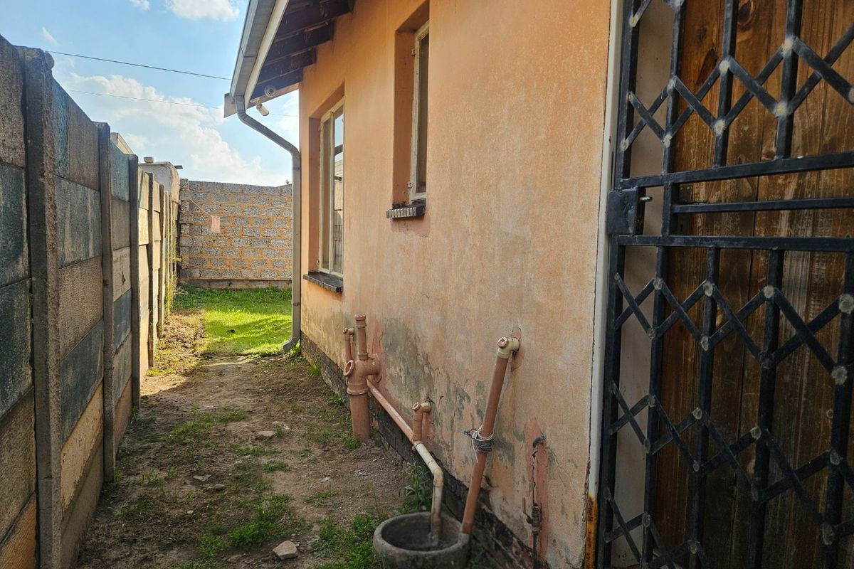 Rear exterior view of the property showing weathered wall finishes, exposed plumbing, a concrete drainage basin, and a metal security gate.