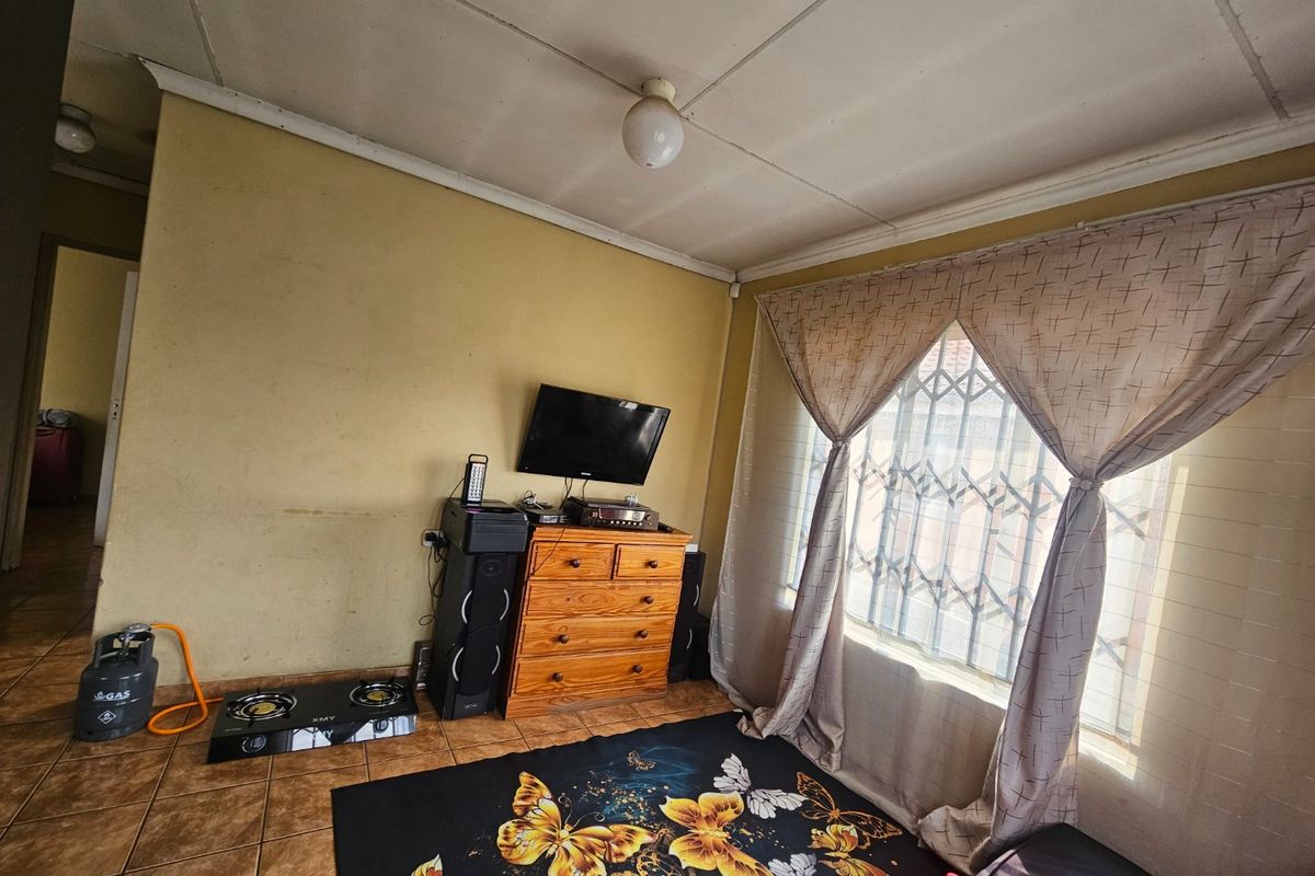 A living room featuring tiled flooring, a window with a security gate, and standard ceiling tiles, requiring cosmetic updates to improve the overall aesthetic.