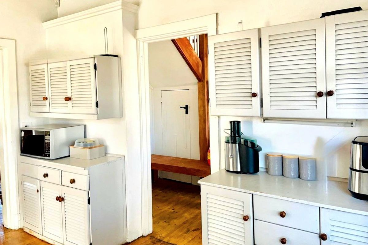 A dated kitchen area featuring white louvered cabinetry and timber flooring, with a doorway leading to an adjacent hallway space.