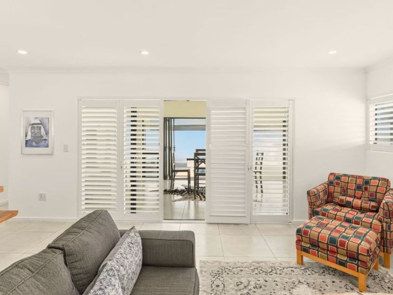 Bright living room featuring neutral tiled flooring, recessed downlights, and white plantation shutters opening onto an outdoor patio area.