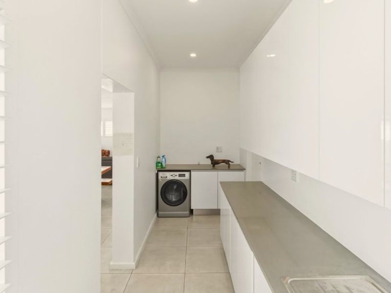 A clean and modern laundry room featuring white high-gloss cabinetry, stone countertops, a stainless steel sink, and neutral floor tiles.