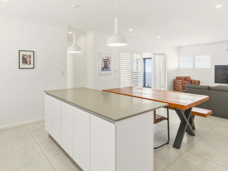 Modern open-plan living area featuring a sleek kitchen island with stone countertops, tiled flooring, and plantation shutters leading to an outdoor space.