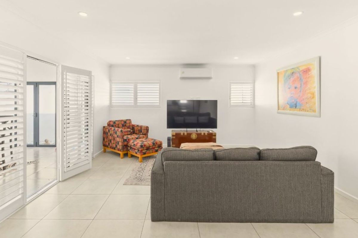 A bright and spacious living room featuring neutral tiled flooring, recessed downlights, plantation shutters, and direct access to an outdoor area via sliding glass doors.