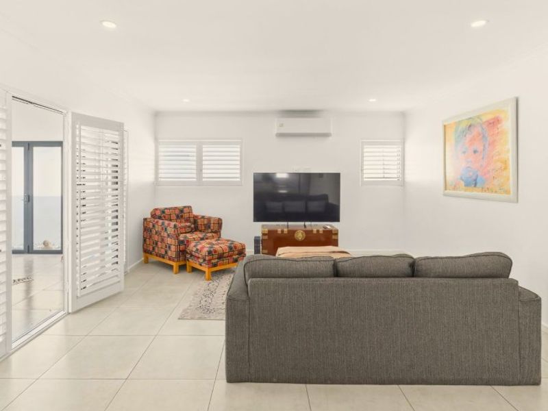 A bright and spacious living room featuring neutral tiled flooring, recessed downlights, plantation shutters, and direct access to an outdoor area via sliding glass doors.