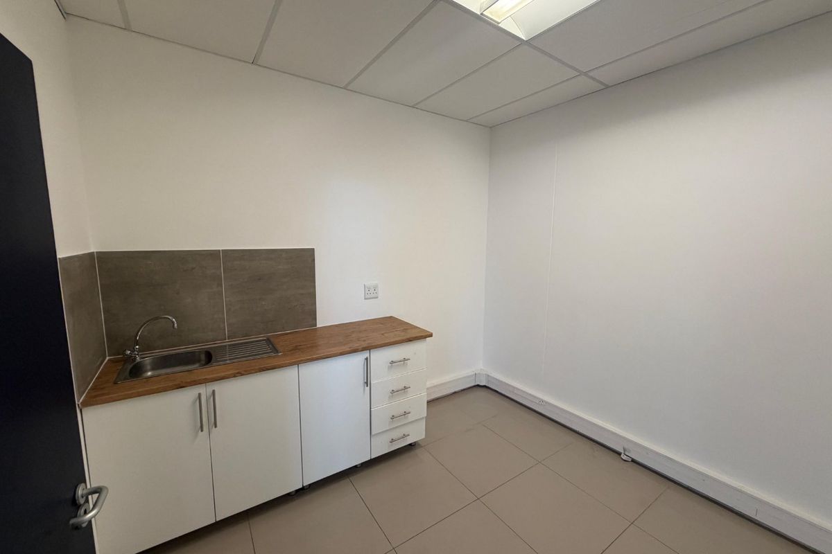 A clean, empty kitchenette featuring white cabinetry, a stainless steel sink, wood-look laminate countertop, and neutral tiled flooring in a commercial office space.