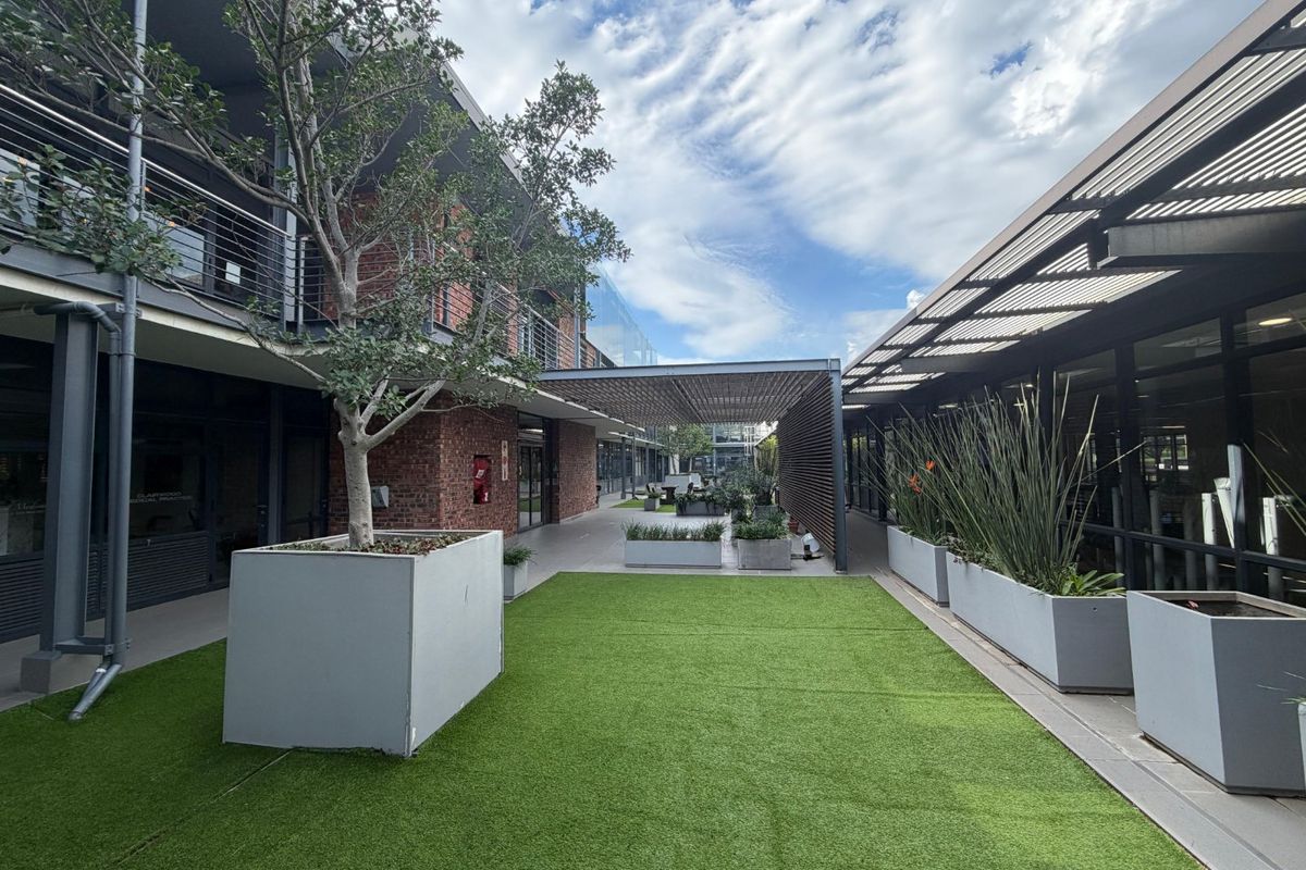 A modern commercial courtyard featuring exposed brick architecture, a metal pergola, artificial turf, and integrated concrete planters between office building wings.