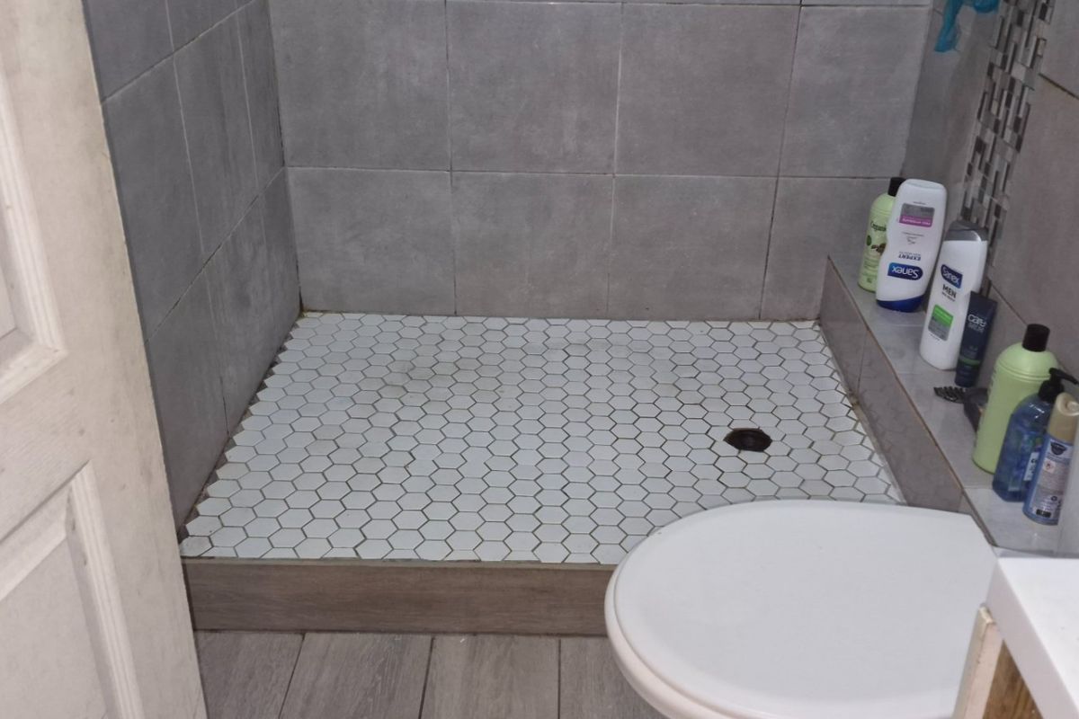 A functional bathroom featuring a walk-in shower with hexagonal floor tiles, wall tiling, a standard toilet, and a vanity basin.