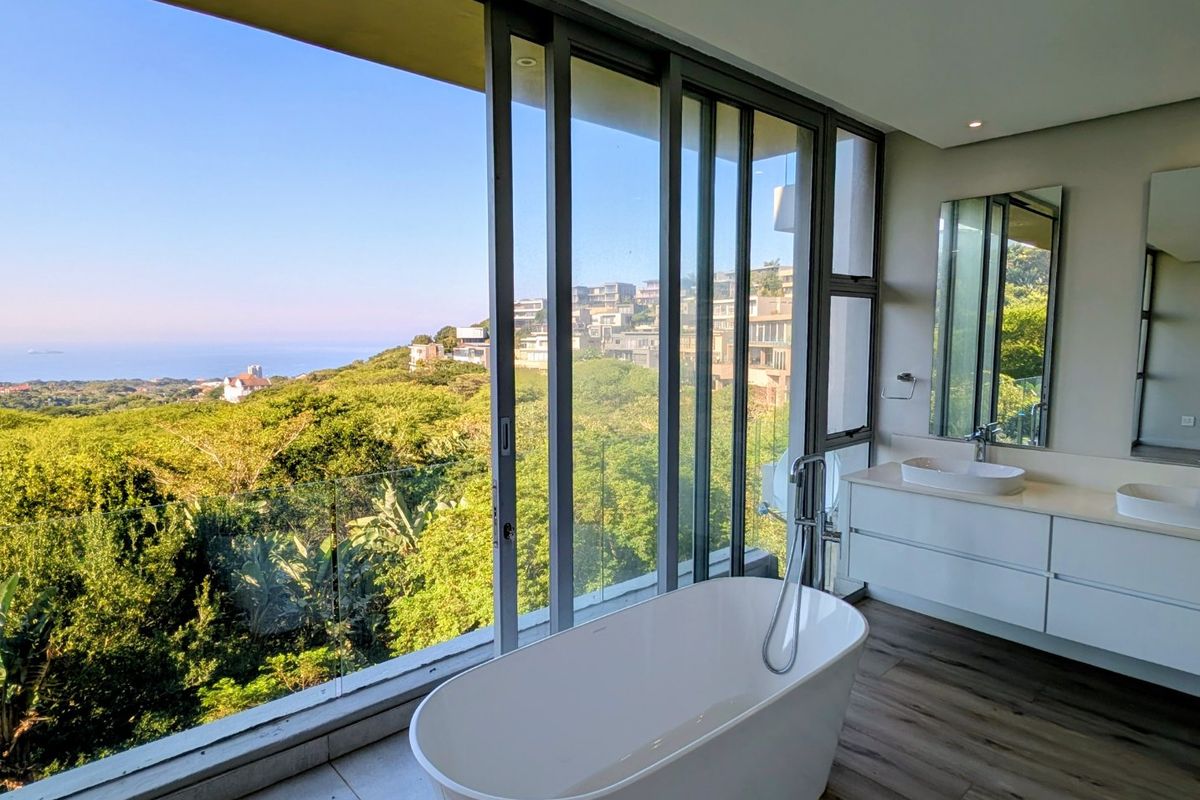Modern bathroom featuring a freestanding bathtub, double vanity with vessel sinks, and expansive floor-to-ceiling glass doors offering panoramic coastal and hillside views.