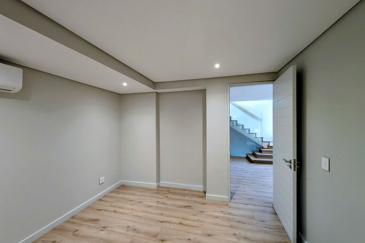 A clean, modern, and empty bedroom featuring light timber-look flooring, recessed downlighting, and a wall-mounted air conditioning unit.