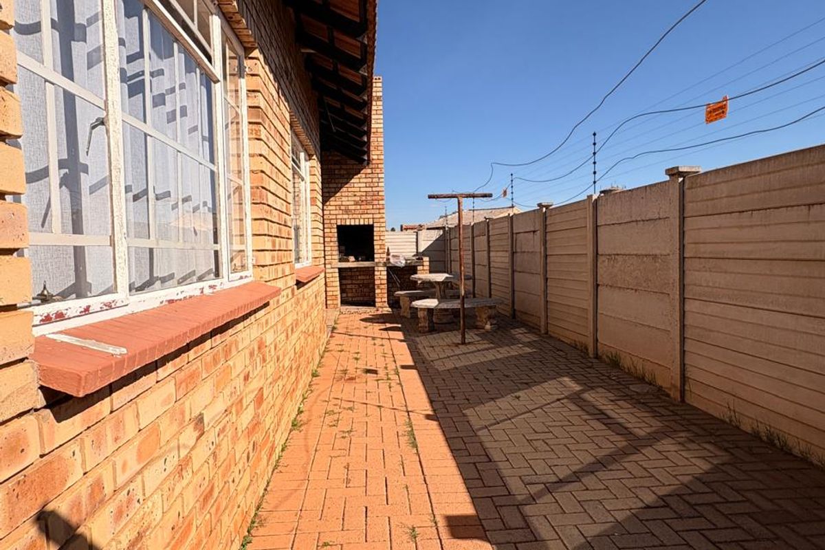 A side exterior view of the property featuring a brick facade, paved walkway, built-in braai area, and secure precast boundary walling.