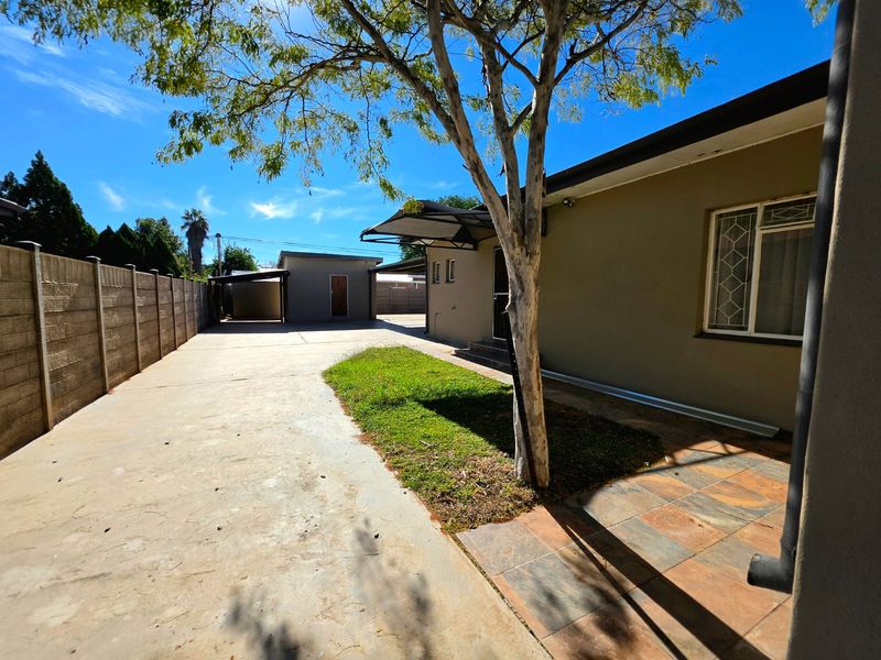Rear exterior view featuring a spacious concrete driveway, precast boundary wall, tiled patio area, and a detached carport structure.