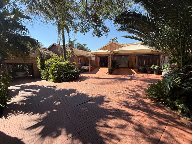 A spacious front exterior featuring extensive brick paving, a covered carport area, and established garden landscaping with mature trees.
