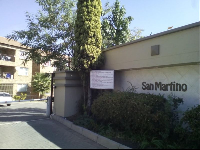 Entrance view of the San Martino complex featuring a security boom gate, perimeter wall, and paved driveway access.