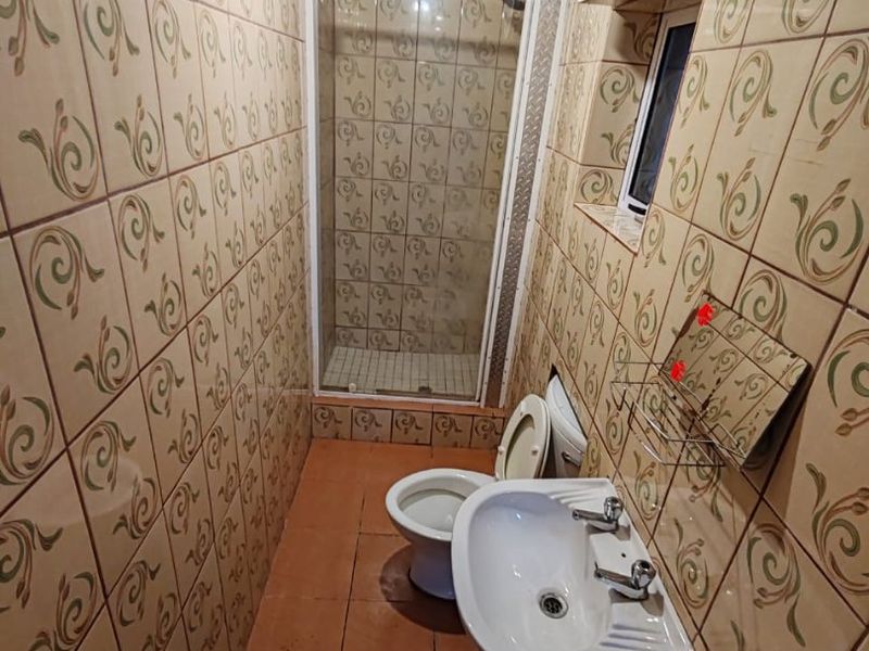 A compact bathroom featuring a shower enclosure, pedestal basin, and toilet, finished with dated patterned wall tiles and a timber ceiling.