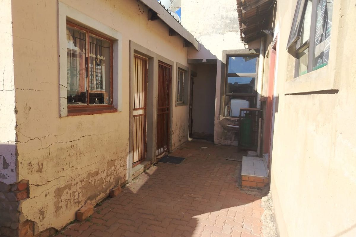 A narrow exterior rear passage featuring paved flooring, security gates, and visible signs of exterior wall deterioration and plaster cracking.