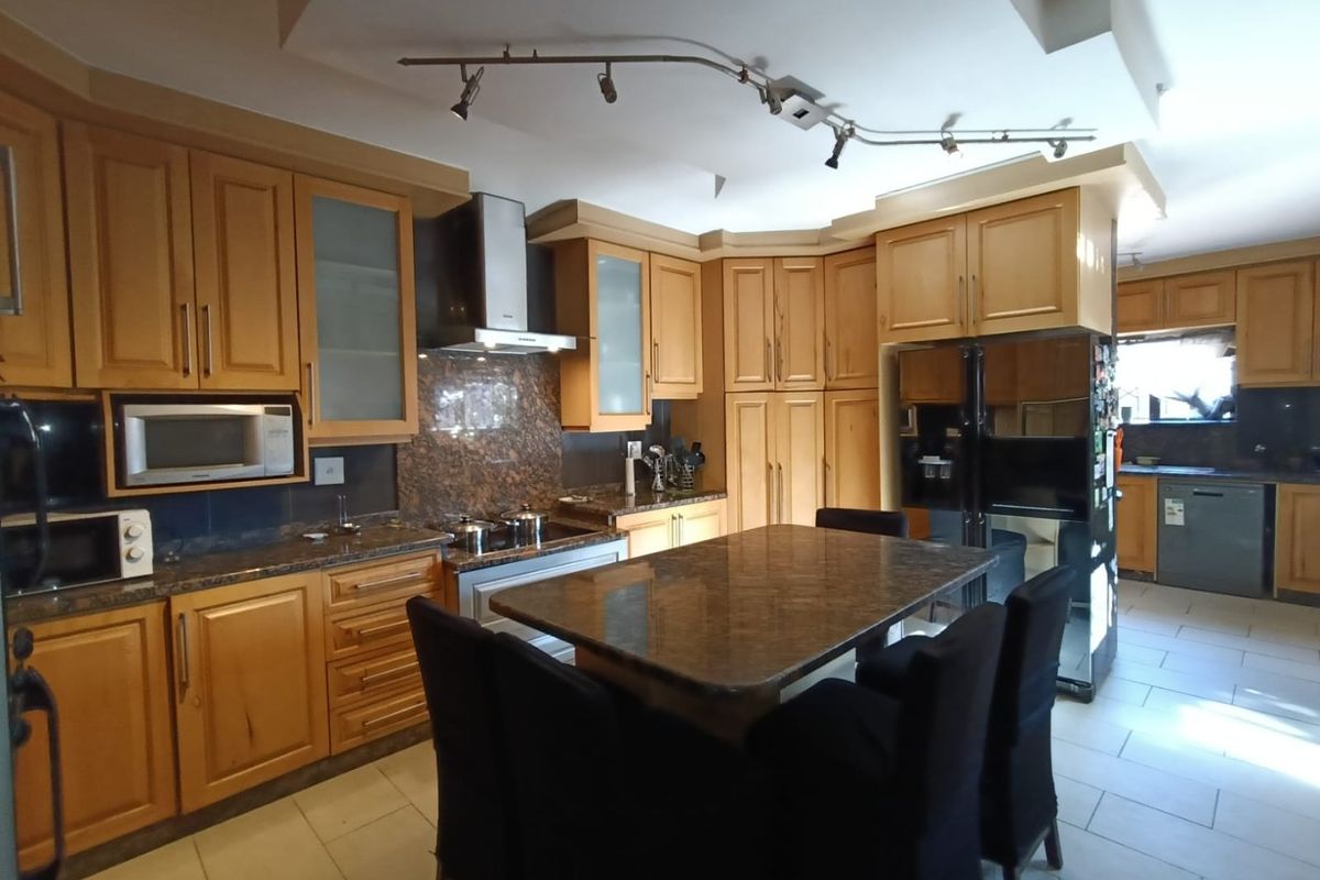 A spacious kitchen featuring wooden cabinetry, granite countertops, integrated appliances, and a central island, with an adjoining scullery area.