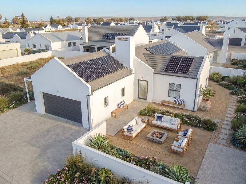 Aerial view of a modern white house featuring solar panels, a paved driveway, a garage, and a landscaped courtyard with a fire pit area.