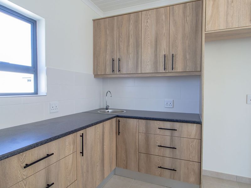 A clean, modern scullery featuring light wood-grain cabinetry, dark laminate countertops, a stainless steel sink, and white tiled splashbacks.