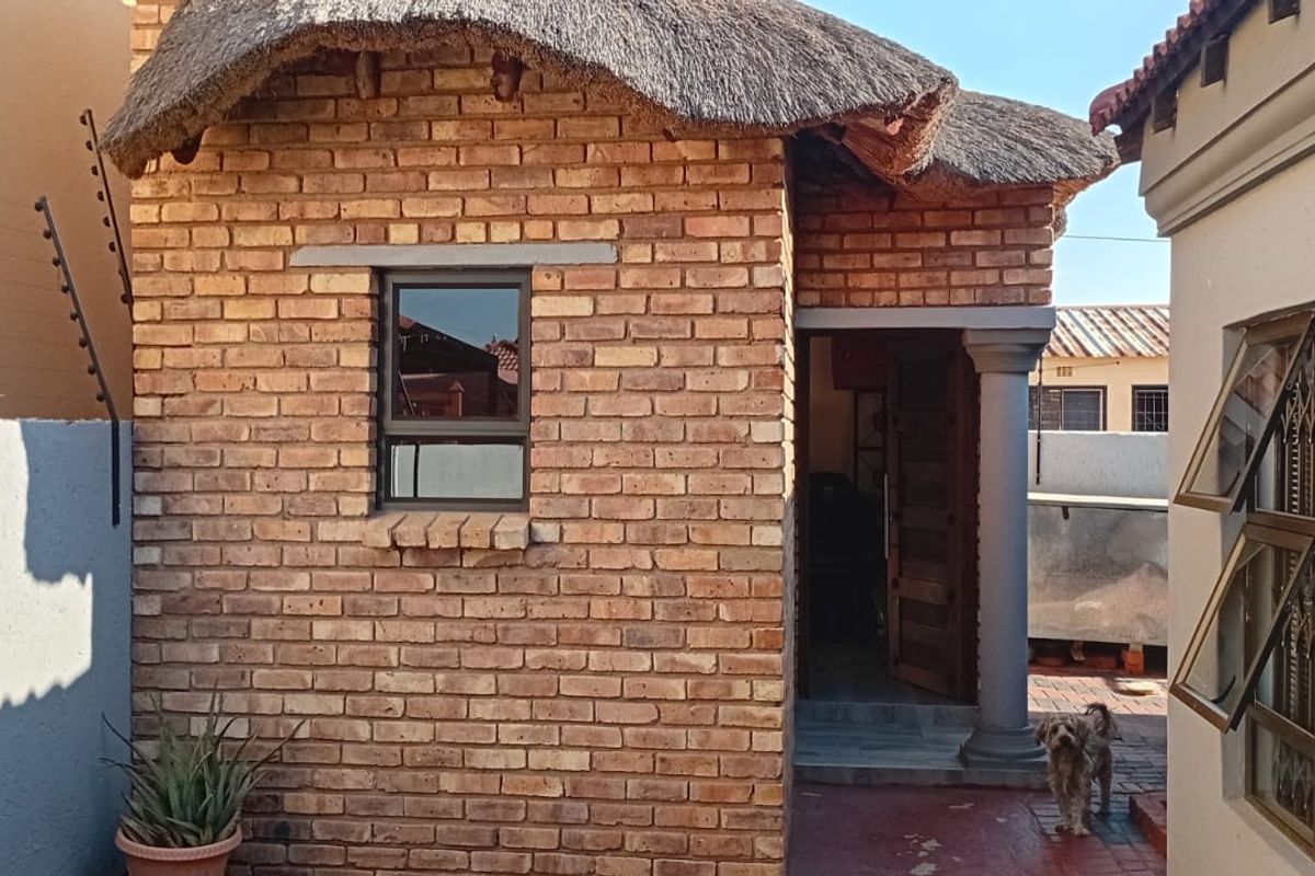 A rear exterior view of a face brick structure featuring a traditional thatch roof, concrete column, and security fencing.