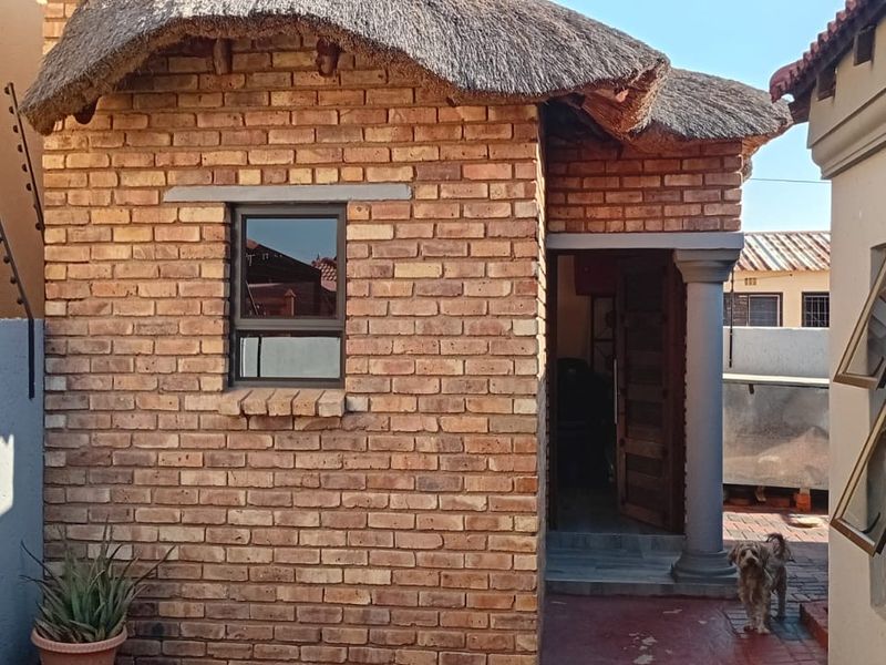 A rear exterior view of a face brick structure featuring a traditional thatch roof, concrete column, and security fencing.