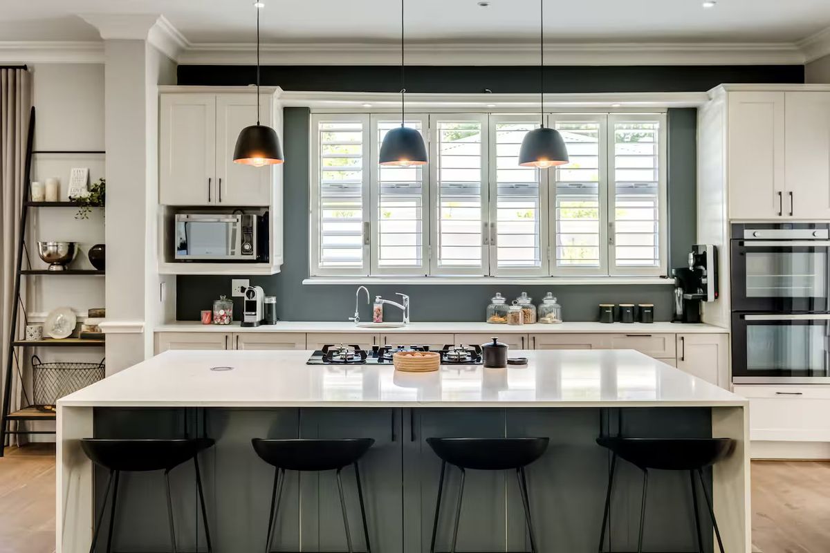 Modern kitchen featuring a large central island with quartz countertops, gas cooktop, built-in double oven, and elegant shaker cabinetry with plantation shutters.