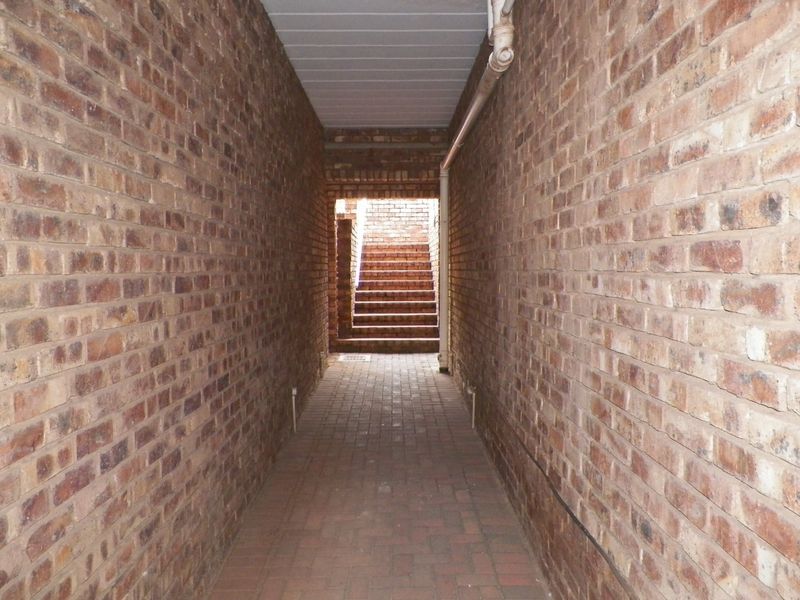 A narrow outdoor brick-paved walkway leading to an external staircase, featuring exposed brick walls and visible utility piping.