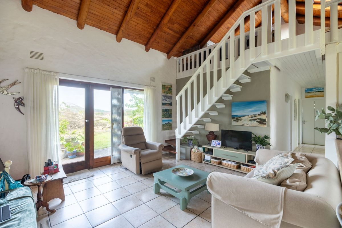 A bright living room featuring a high exposed timber beam ceiling, tiled flooring, and a staircase leading to a mezzanine level with garden access.