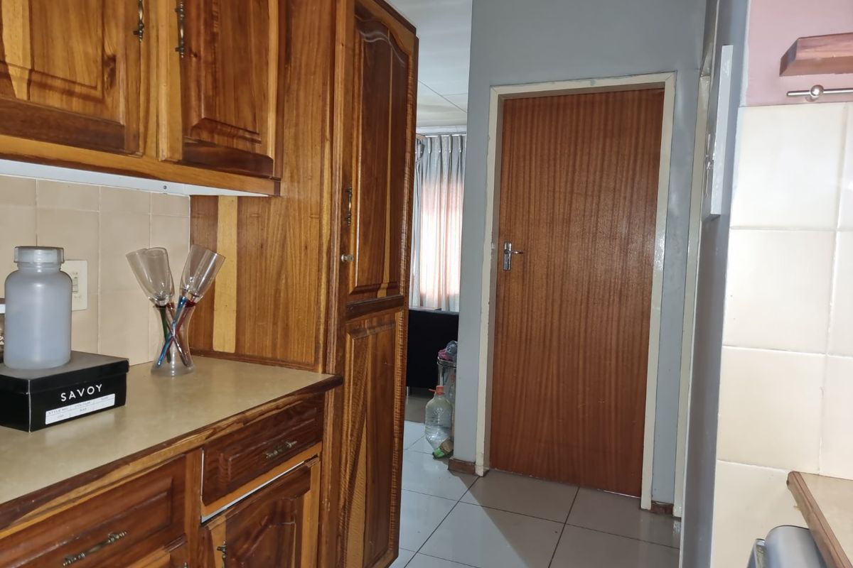 A kitchen area featuring traditional wooden cabinetry, tiled backsplash, and tiled flooring, leading to an adjacent room through a wooden door.