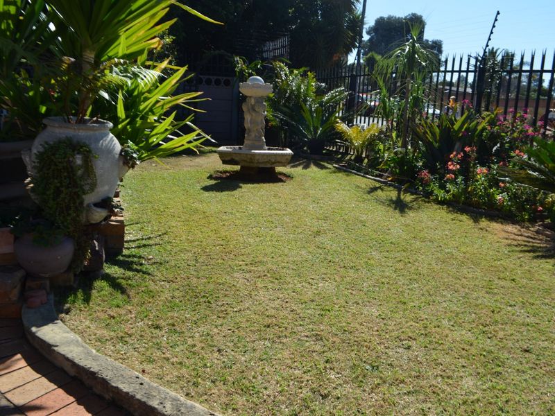 A well-maintained residential garden featuring a central stone fountain, established landscaping, and secure metal perimeter fencing.