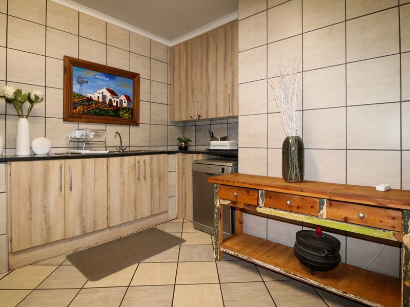 A functional scullery area featuring light wood-grain cabinetry, dark countertops, a stainless steel sink, and full-height wall tiling.