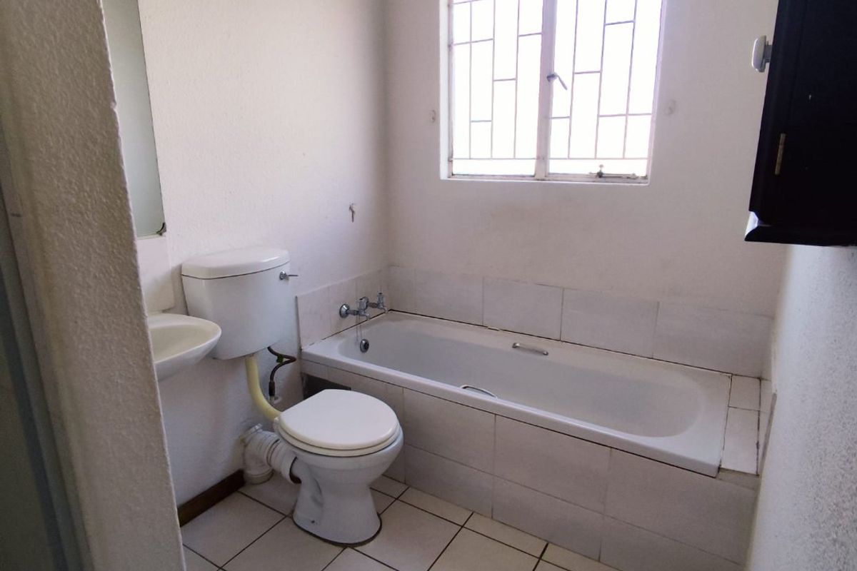 A compact bathroom featuring a standard bathtub, toilet, and wall-mounted basin with tiled flooring and security bars on the window.