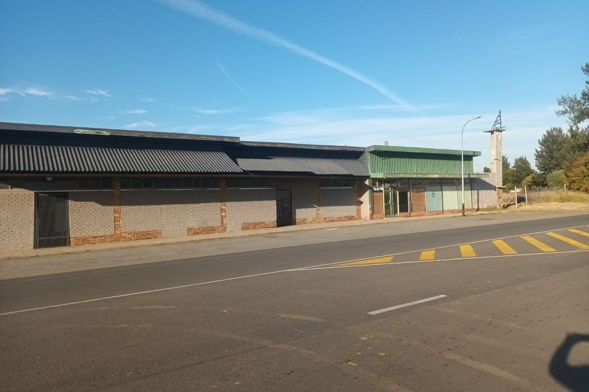 Commercial property frontage featuring a long brick facade, corrugated metal roofing, and large storefront windows along a main road.