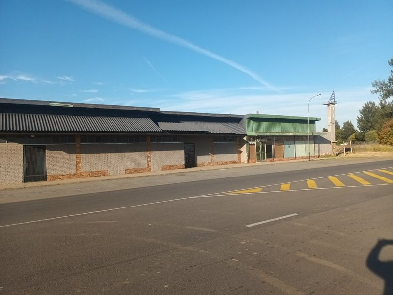 Commercial property frontage featuring a long brick facade, corrugated metal roofing, and large storefront windows along a main road.