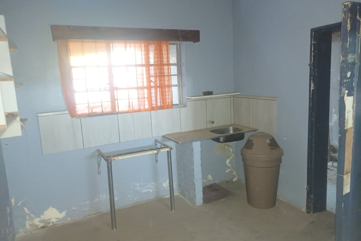 A basic, empty kitchen area featuring a small stainless steel sink, tiled splashback, and security bars on the window, requiring significant cosmetic refurbishment.