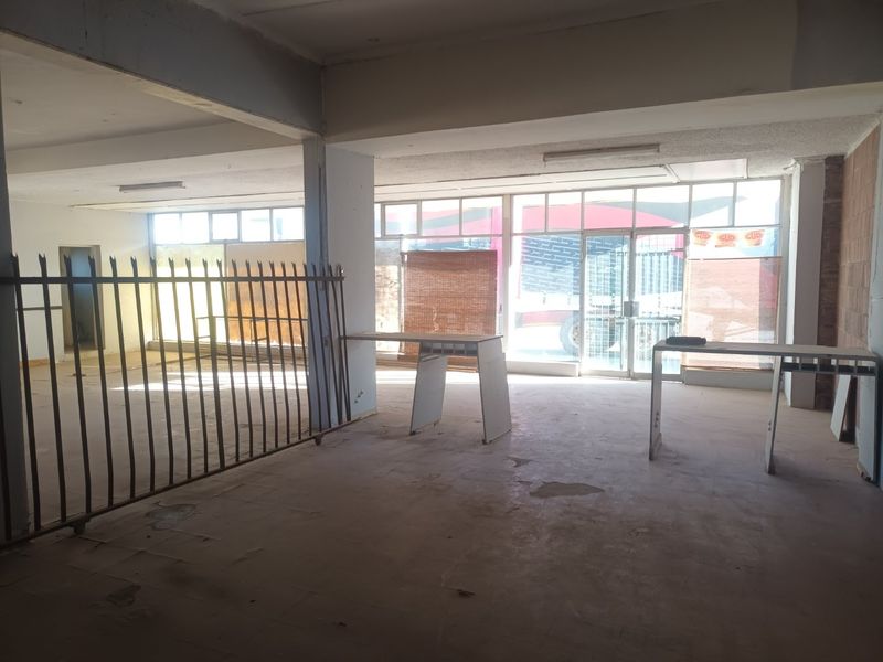Spacious open-plan commercial unit featuring large glass storefront windows, tiled flooring, and a metal security gate, suitable for retail or office use.