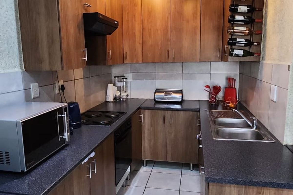 A functional galley-style kitchen featuring wood-finish cabinetry, dark laminate countertops, a double basin sink, and integrated cooking appliances with modern pendant lighting.