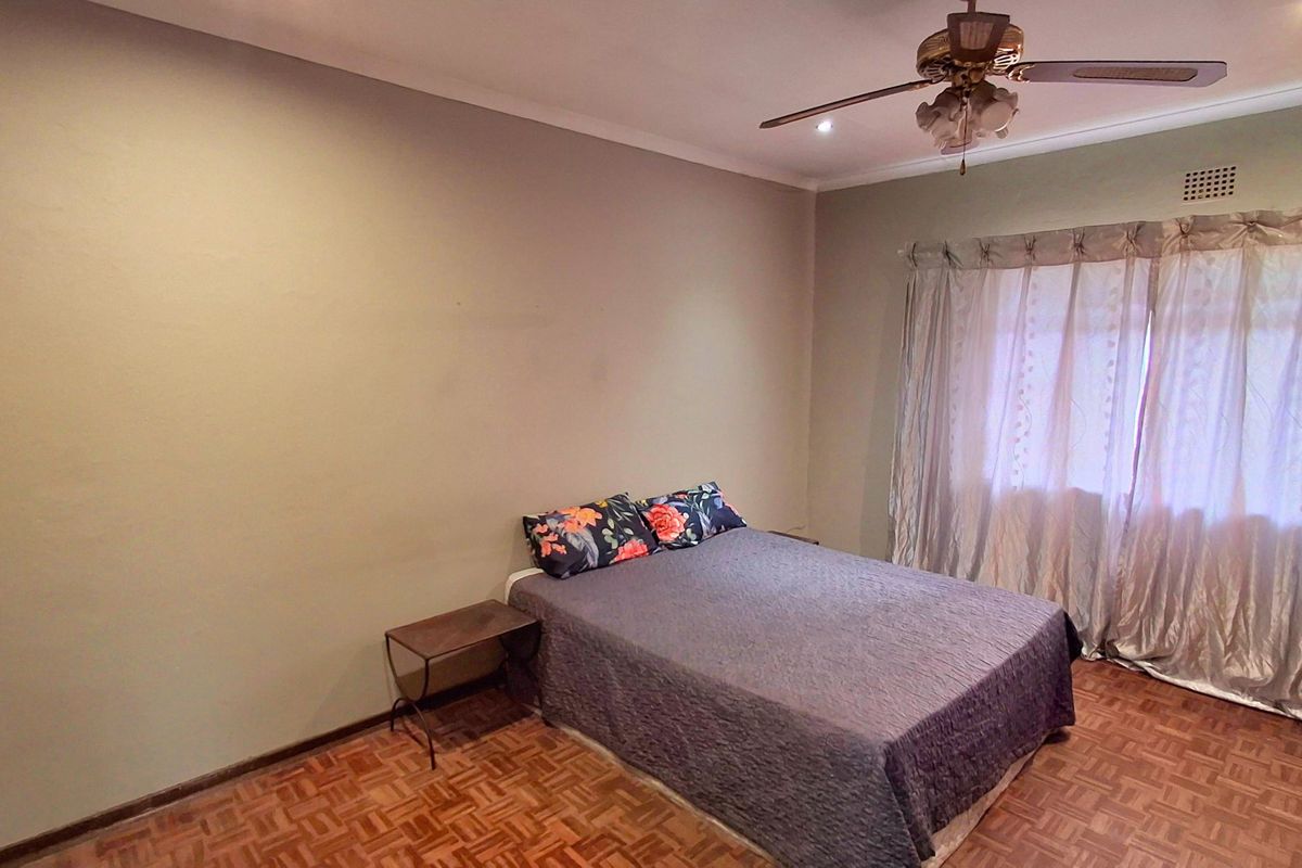 A spacious bedroom featuring classic parquet flooring, a ceiling fan, and large windows with curtains, offering a neutral canvas for personalization.