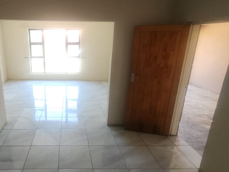 An empty living area featuring light-colored floor tiles, a wooden door, and natural light from a large window.
