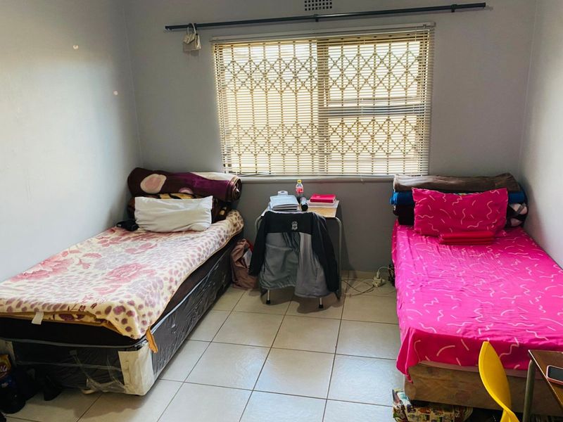 A functional bedroom featuring tiled flooring, a window with security bars, and venetian blinds, providing a practical space for rest.