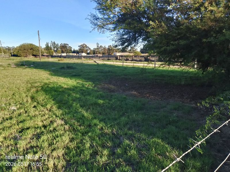 A view of a vacant residential plot featuring grassy terrain, mature trees, and perimeter wire fencing.