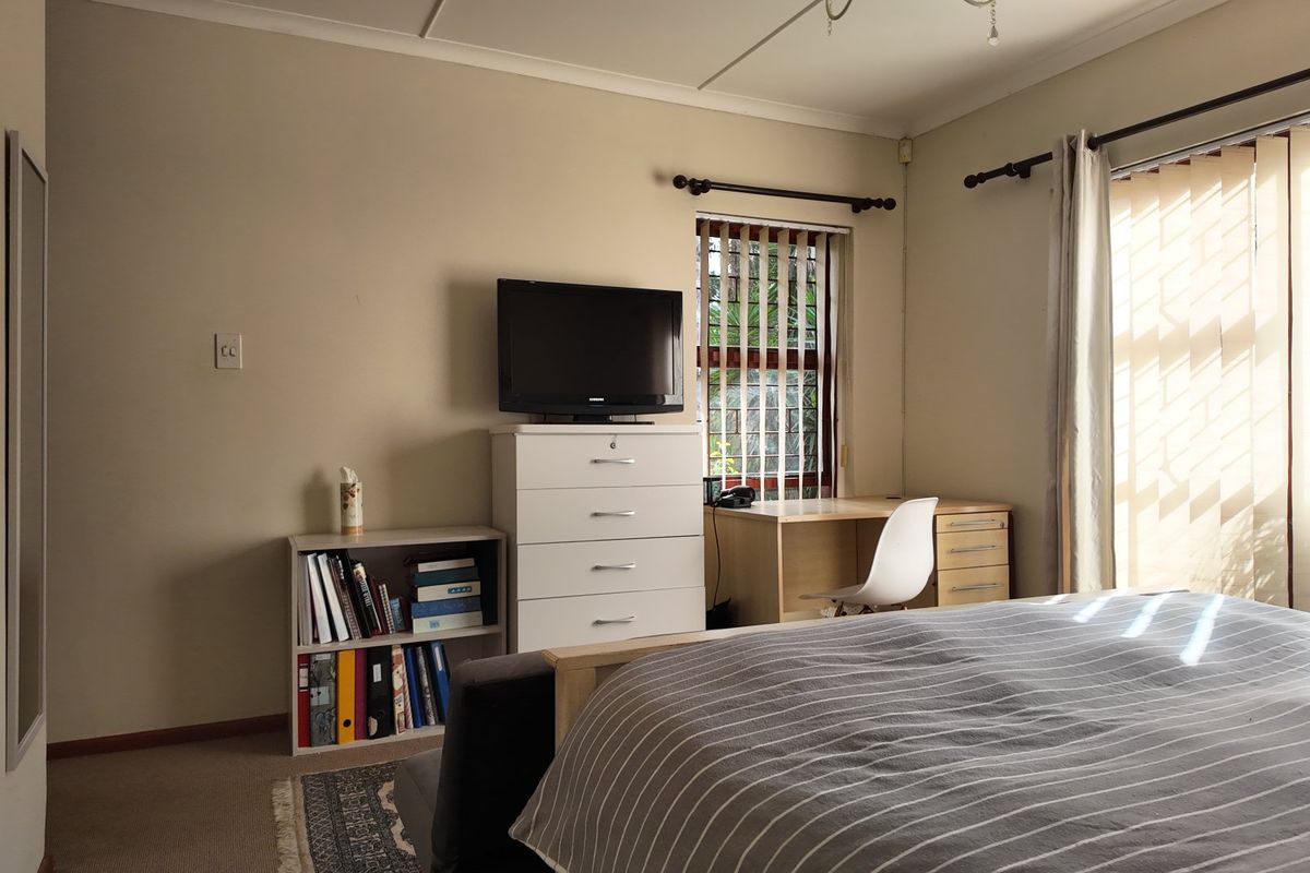 A functional bedroom featuring neutral wall tones, carpeted flooring, and a large window with vertical blinds allowing natural light.