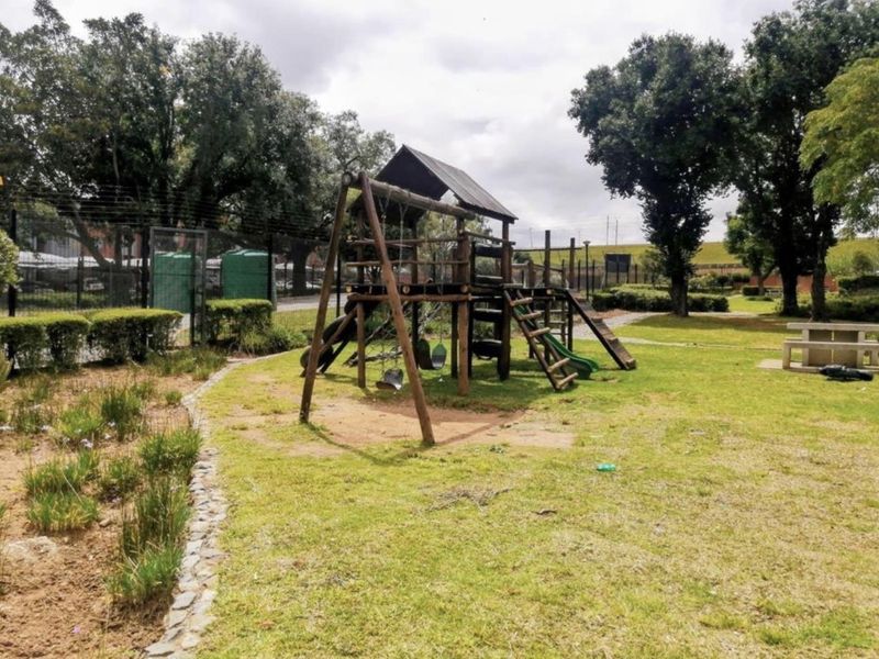 A communal garden area featuring a wooden playground structure, stone picnic table, and landscaped garden beds within a fenced residential complex.
