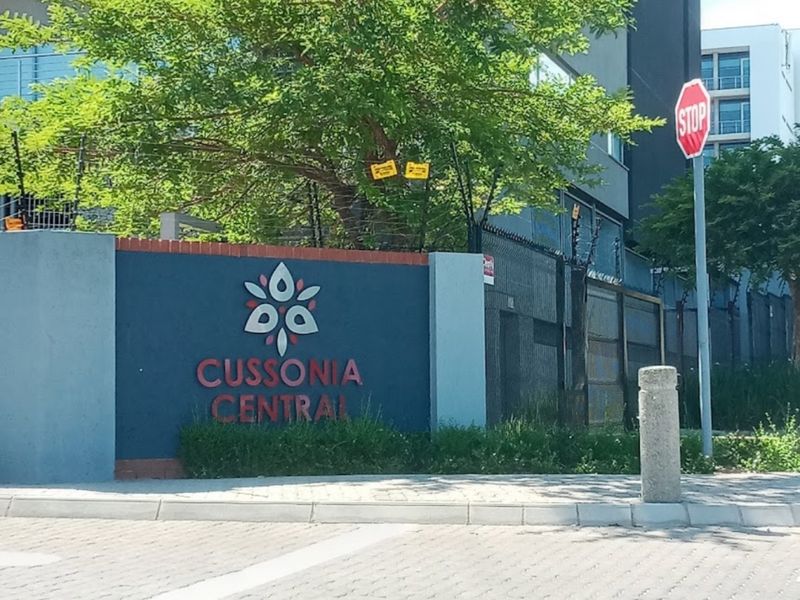 Street view of the Cussonia Central apartment complex entrance, featuring paved access and secure perimeter fencing.