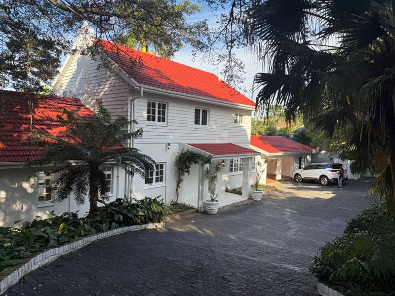 A charming two-story home featuring white weatherboard cladding, a vibrant red tiled roof, and a spacious paved driveway leading to a covered entrance.
