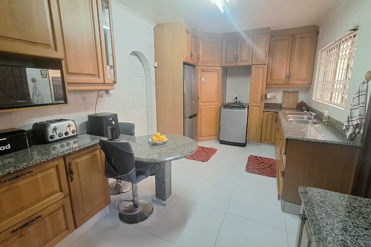 A functional kitchen featuring wooden cabinetry, granite countertops, a double basin sink, and tiled flooring with an integrated microwave and laundry appliance space.