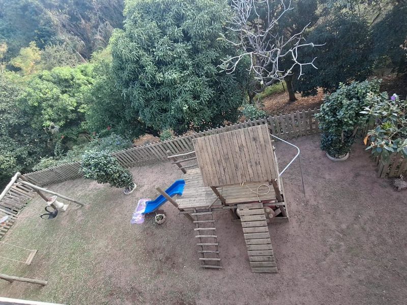 An elevated view of a backyard garden featuring a wooden play structure, slide, and perimeter fencing surrounded by mature trees.