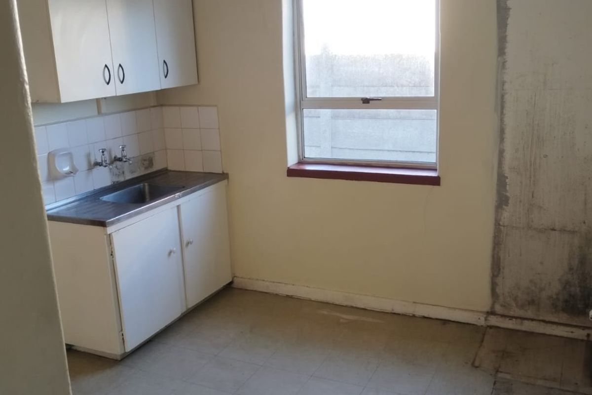 A small, dated kitchen area featuring basic cabinetry, a stainless steel sink, and significant wear to the flooring and wall finishes.
