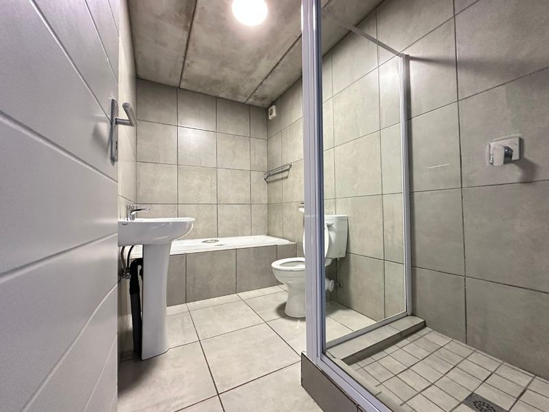 Modern bathroom featuring a pedestal basin, built-in bath, and separate shower enclosure with neutral floor-to-ceiling tiling and an industrial-style exposed concrete ceiling.